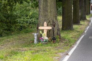 Shows a light wood roadside cross with flowers and gifts around it