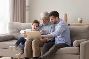 Father, son, and grandson sitting on a couch, looking at a laptop together while smiling