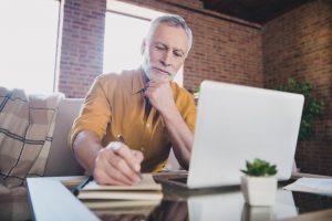 Older man sitting on couch with laptop sitting on coffee table in front of him, thinking and writing on pad of paper