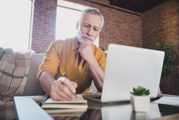 Older man sitting on couch with laptop sitting on coffee table in front of him, thinking and writing on pad of paper