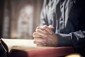 Person sitting quietly with religious book, hands clasped in prayer