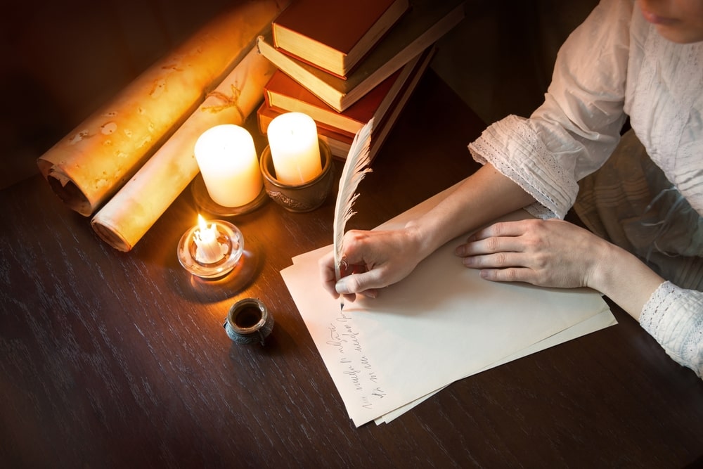 Waman in white dress sitting at table with candles, paper, and quill pen