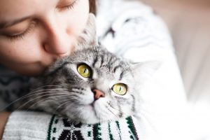 Young woman holds gray, striped cat in her arms and kisses the cat's head