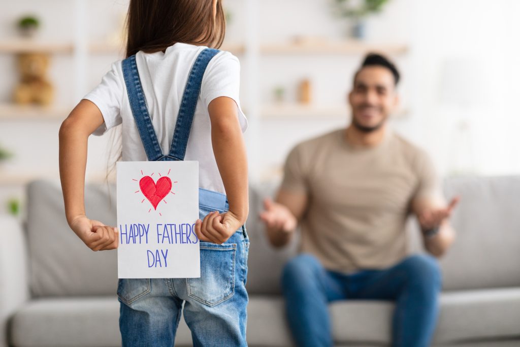 Rear view of little girl holding paper card behind her back, greeting positive adult man with Father's Day.