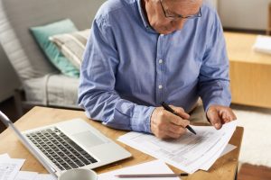 Older man in blue button-down shirt sitting at table at home, completing forms