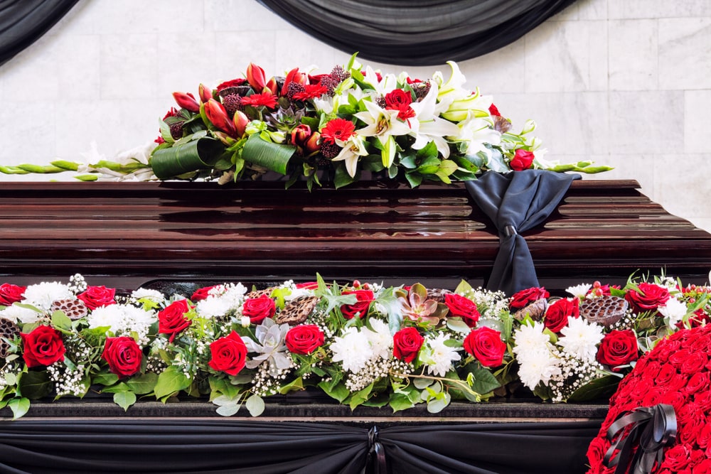 Casket surrounded by floral arrangements, including floral scarf of red and white flowers