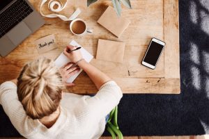 View from above of woman sitting at table writing a thank you note with coffee and cell phone nearby