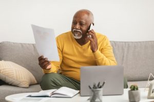 Older man in yellow shirt sitting on his couch as he talks on phone and looks at form