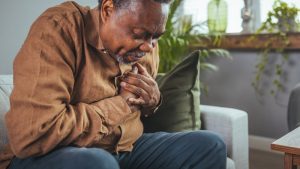 Man sitting on couch at home, hand on chest as he experiences chest pain that could be broken heart syndrome