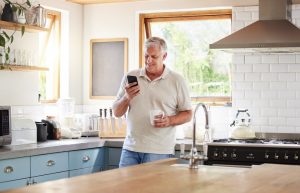 Older man standing in his kitchen at home, reading messages on his phone