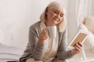 Mature woman sitting at home, drinking a cup of coffee and holding a picture frame as she remembers a lost loved one