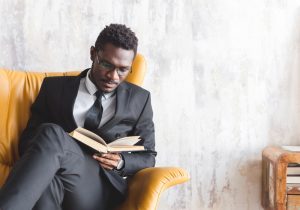 Young man dressed in black suit, sitting in a yellow chair as he reads and contemplates a book