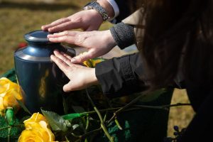 Blue urn surrounded by yellow flowers with three different people reaching out to the touch top gently in remembrance