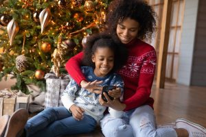Mother and young daughter sitting in front of Christmas tree as they watch something funny on a smartphone
