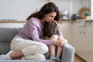 Mother and son sitting on couch at home, mother hugging son close as he cries and is upset