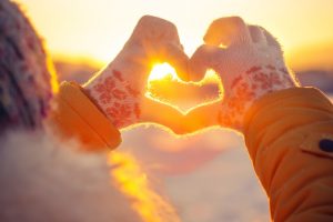 Person bundled up in coat, winter hat, and gloves, standing outside, creating heart with hands and the sunset shining through the heart shape