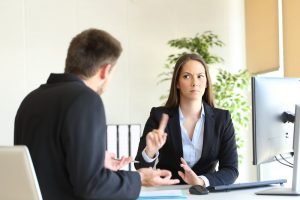 Unsympathetic professional woman sitting at desk across from client with upset look on her face and holding up index finger sternly