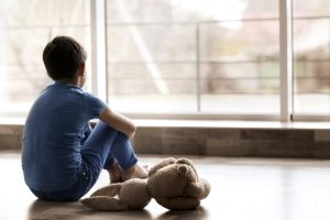 Young boy sitting by himself, looking out a window with a stuffed animal beside him