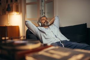 Mature man in casual shirt reclining on couch as he listens to music on headphones