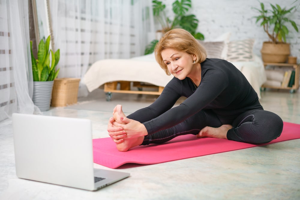 Mature woman sitting on a pink exercise mat, stretching her right leg out in front of her