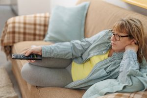 Middle-aged woman sitting on couch at home, mindlessly choosing something to watch