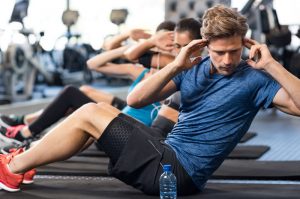 Young man in group class, sitting on a black exercise mat and doing an ab exercise