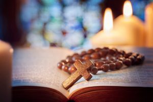 Wooden rosary laying on top of open Bible with lit candles in background
