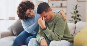 Husband and wife sitting at couch at home; husband sad and wife comforting him