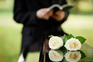 Two white roses sitting on casket with clergy person in background holding an open book