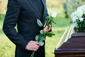 Man in black suit holding white rose as he stands next to casket