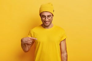 man wearing all yellow standing in front of a yellow background pointing at his shirt