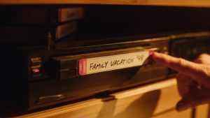 Person pushing an old home video tape labeled "Family Vacation '98" into a VCR