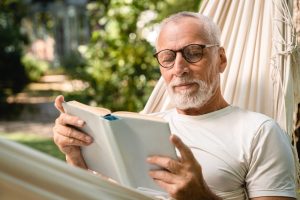 Mature man with glasses and beard sitting in a hammock outside and reading a book of poems