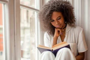 Young woman sitting beside window as she reads a book of poems