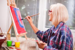 older woman in a plaid shirt painting a canvas on a tabletop easel