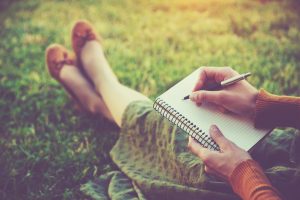 woman in a green skirt with brown shoes writing in a notebook while sitting outside on grass