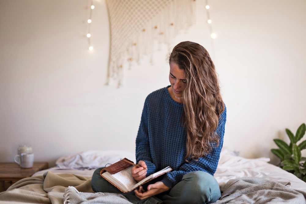 Woman in blue sweater sitting on bed, writing in journal