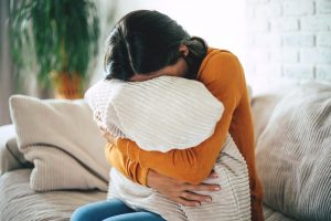 Woman with black hair sitting on couch, hugging a pillow close and pressing her face into it