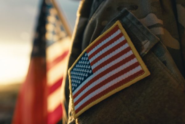 Close up view of a veteran's arm in uniform with American flag badge and flag in the background