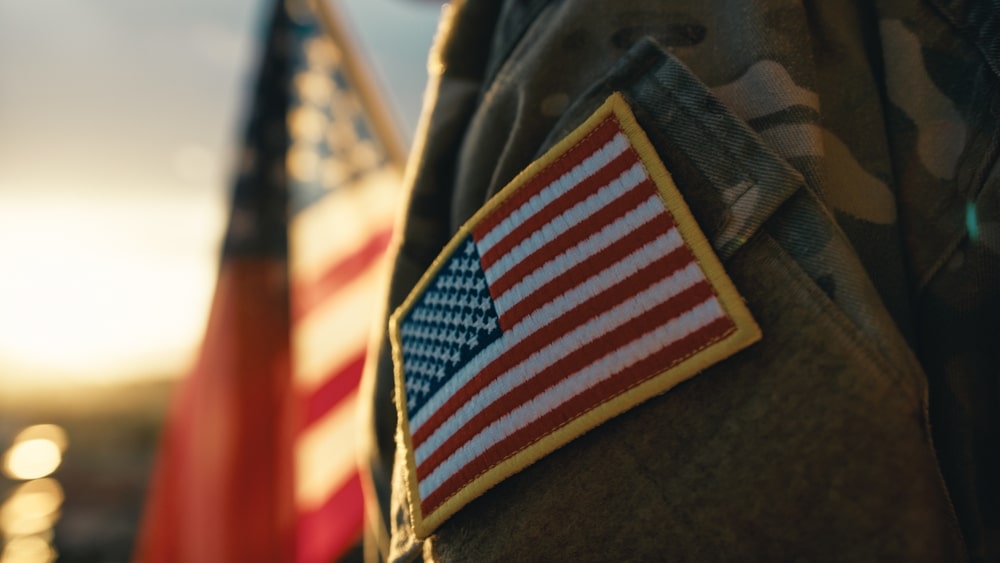 Close up view of a veteran's arm in uniform with American flag badge and flag in the background