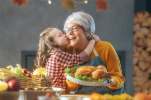 Grandmother and granddaughter in the kitchen; grandmother holding platter with turkey and smiling as granddaughter kisses her cheek