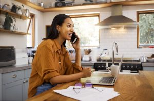 Woman sitting in her kitchen, talking on the phone with laptop on table in front of her