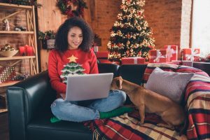 Young woman sitting on couch at home, wearing an ugly Christmas sweater and working on her laptop
