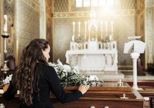 Woman with long brown hair standing next to a casket, placing her hand tenderly on the top of it