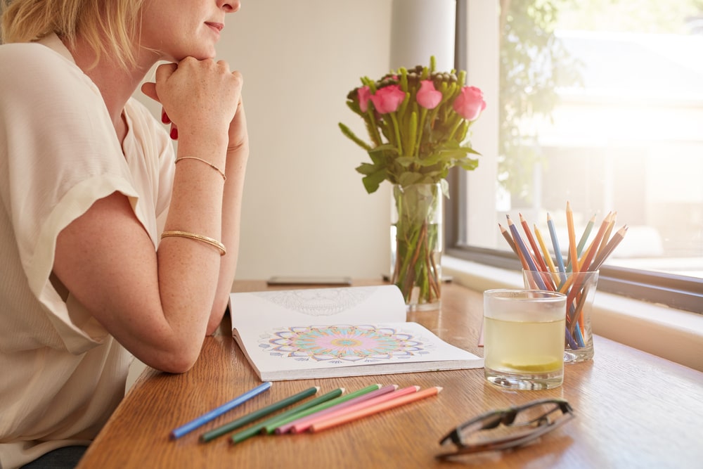 Woman sitting at a table at home with flowers, colored pencils, and a coloring book in front of her