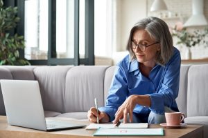 Mature woman in blue shirt sitting on couch, working on laptop and writing down notes