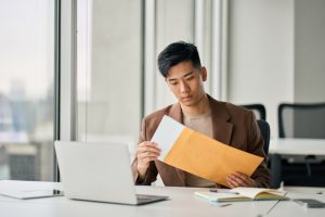 Asian man in brown jacket sitting at a table while opening an envelope with papers inside