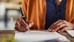 Woman in mustard yellow cardigan sitting at table, signing documents