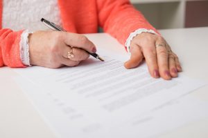 Older woman in bright orange sweater sitting at table, signing a legal document