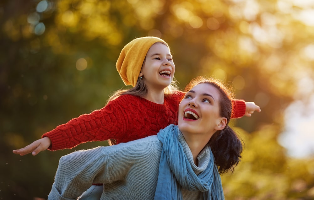 young woman outside holding a little girl on her back who has her arms outstretched like an airplane